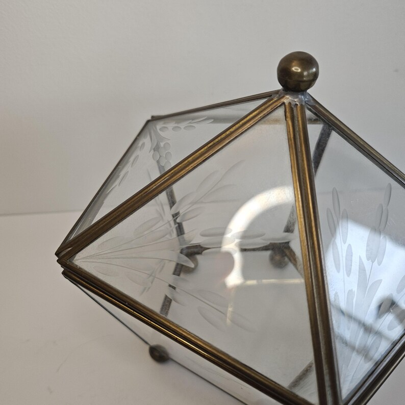 May include: A geometric glass and brass box with a clear glass top and etched floral designs. The box has a brass frame and a brass knob on top.