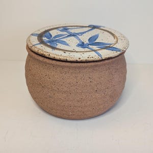 Peut inclure: Un pot en céramique ronde de couleur beige moucheté avec un couvercle assorti. Le couvercle présente une surface crème avec des taches brunes et un motif de feuilles de bambou bleu peint à la main dans un cercle brun. Finition texturée.
