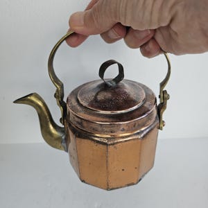 Peut inclure: Petite théière en cuivre avec une anse et un bec en laiton. La théière a un couvercle avec une petite poignée en boucle. Le corps de la théière est de couleur cuivre chaud, et le bec et la poignée sont en laiton.