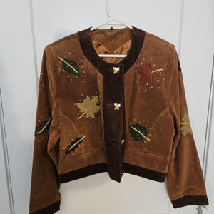 May include: Brown suede jacket with leaf appliqués in various colours. The jacket features a round collar, dark brown trim, and gold-coloured button closures. Small gold-coloured studs are scattered around the leaves.