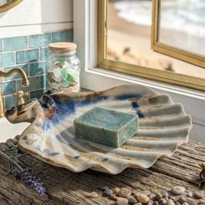 May include: A ceramic soap dish shaped like a large scallop shell with a cream-coloured base, blue accents, and a textured surface. A rectangular bar of sage green soap rests in the centre of the shell, which features deep, radiating ridges.