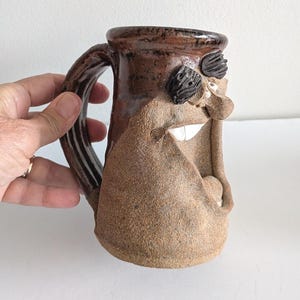 Peut inclure: Une tasse en céramique représentant un visage sculpté avec une large bouche ouverte, un nez proéminent et des sourcils texturés foncés. La tasse présente une partie supérieure vernie brun foncé et une base beige à la texture granuleuse.