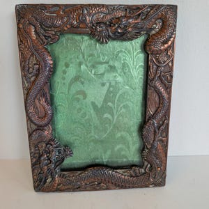 May include: An antique picture frame with a copper finish, featuring detailed dragon carvings. The frame's edges and corners are adorned with dragon designs. The centre holds green fabric with a floral pattern.