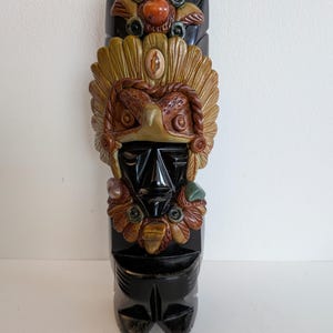 May include: A carved, black wooden totem with a detailed face and headdress. The totem features a central face with a beak-like nose and a feathered headdress in shades of brown and tan. Decorative elements include colorful stone accents.