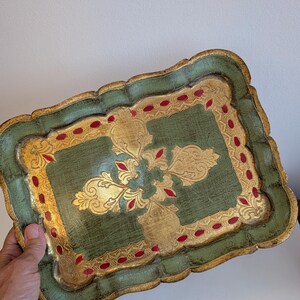 Peut inclure: Un plateau décoratif rectangulaire avec un bord festonné. Le plateau présente un fond vert vieilli avec des accents dorés et un motif floral doré central. Des points rouges et une bordure dorée bordent le plateau.