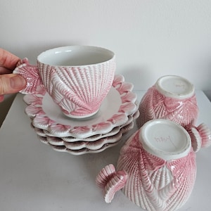 May include: Set of three pink and white porcelain teacups and saucers with a seashell design. The cups and saucers have a raised, textured design that resembles seashells. The cups have a small handle on the side. The saucers have a scalloped edge. The cups and saucers are made in Italy.