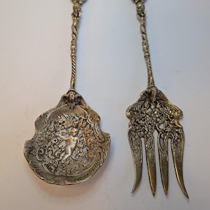May include: An ornate silver serving spoon and fork set. The spoon has a detailed cherub design in the centre, surrounded by floral patterns. The fork has three tines and a matching floral design. Both pieces have decorative handles.
