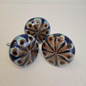 Peut inclure: Trois boutons de meuble en céramique avec un motif de plume de paon. Les boutons sont principalement bleus, avec des accents de marron, de beige et de bleu clair. Chaque bouton a une vis métallique pour l'installation. Les boutons sont décoratifs et conviennent aux meubles.