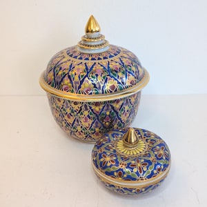 May include: A large lidded bowl and a smaller matching lidded bowl featuring intricate Benjarong style patterns in deep blue, pink, green, and yellow enamel with gold trim. Both bowls have pointed, gold-toned finials on their rounded lids.