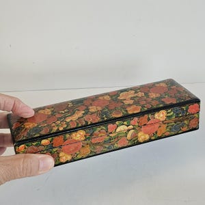 Peut inclure: Une boîte rectangulaire avec un motif floral rouge, orange, jaune et vert sur fond noir. La boîte est tenue par une main. La boîte semble être en bois ou dans un matériau similaire.
