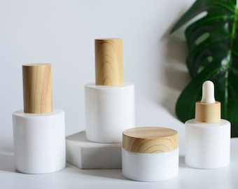 May include: Four white glass bottles with wooden lids. The bottles are different sizes and shapes. The bottles are on a white surface with a green leaf in the background.