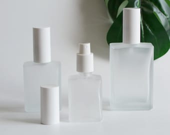 May include: Three frosted glass bottles with white caps. The bottles are different sizes and have a square shape. The bottles are on a white surface.