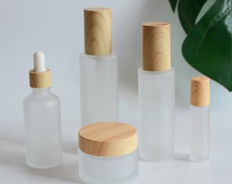 May include: Set of five frosted glass bottles with wooden lids. The bottles are various sizes and shapes, including a dropper bottle, a pump bottle, a jar, and two spray bottles.