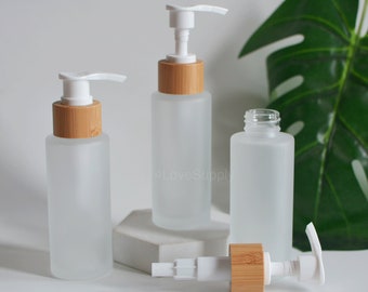 May include: Three frosted glass bottles with bamboo lids and white pump tops. The bottles are on a white surface with a leaf in the background.