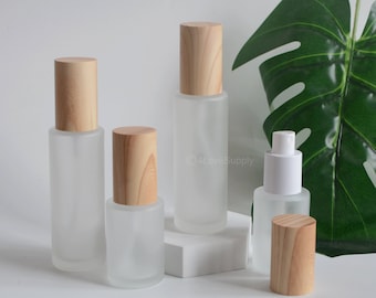 May include: Four frosted glass bottles with wooden caps and a white pump top. The bottles are arranged on a white surface with a green leaf in the background.