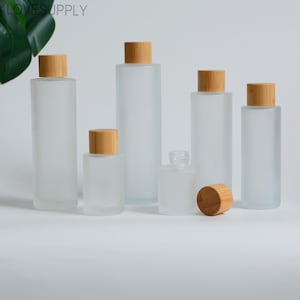 May include: A selection of frosted glass bottles with natural bamboo lids. The bottles are in various sizes and shapes, including a small, round bottle and several taller, cylindrical ones. One lid is separate. The bottles are on a white surface.