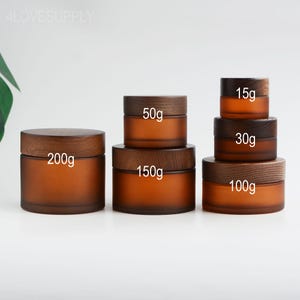 Puede incluir: Una colección de frascos de vidrio color ámbar con tapas de madera. Los frascos están etiquetados con pesos que van desde 15g hasta 200g. Los frascos están dispuestos en una pila, mostrando diferentes tamaños para cosméticos o almacenamiento.