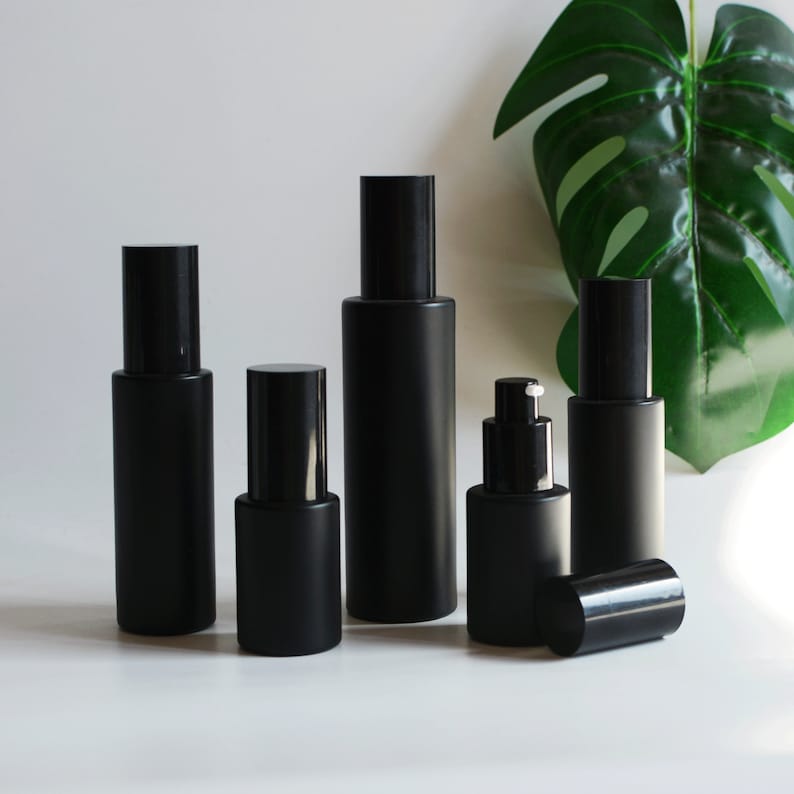 Puede incluir: Conjunto de seis botellas de vidrio negro esmerilado con diferentes tipos de tapas. Las botellas est&aacute;n dispuestas sobre una superficie blanca con una hoja verde en el fondo.
