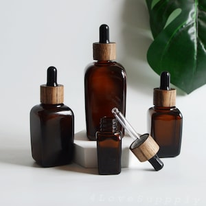 Puede incluir: Tres botellas cuentagotas de vidrio ámbar con tapas de madera y goteros negros. Las botellas son de forma cuadrada y están dispuestas sobre una superficie blanca.