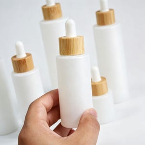 Puede incluir: Una colección de frascos cuentagotas de vidrio blanco esmerilado con collares de bambú marrón claro y tapas de goma blanca. Los frascos son cilíndricos con un acabado mate y liso, diseñados para el almacenamiento de líquidos.