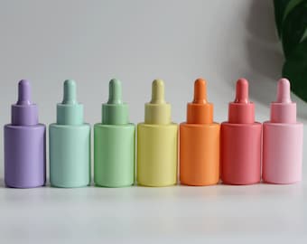 May include: Seven glass dropper bottles in a rainbow of pastel colors, from purple to pink. The bottles are lined up in a row on a white surface.