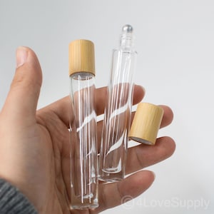 May include: Two clear glass roller bottles with bamboo lids. One bottle has a silver rollerball top. The other bottle has a bamboo lid. The bottles are held in a hand against a white background.
