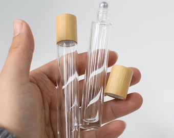 May include: Two clear glass roller bottles with bamboo lids. One bottle has a silver rollerball top. The other bottle has a bamboo lid. The bottles are held in a hand against a white background.