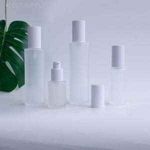 May include: A collection of frosted glass bottles with white caps and a pump dispenser. The bottles vary in size and shape, suitable for storing liquids. The background is a soft white, with a large green leaf in the corner.