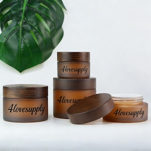 Pode incluir: Vários frascos de vidro âmbar com tampas de madeira são exibidos em uma superfície branca. Os frascos, de tamanhos diferentes, têm a inscrição "4lovesupply". Uma tampa é removida, revelando o conteúdo. Uma grande folha verde está ao fundo.