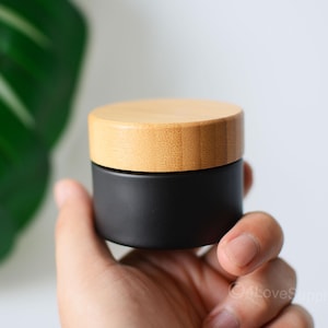 May include: A black glass jar with a bamboo lid. The jar is being held in a hand.