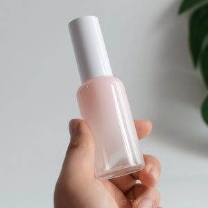May include: A clear glass bottle with a white cap and a light pink liquid inside. The bottle is being held in a hand.