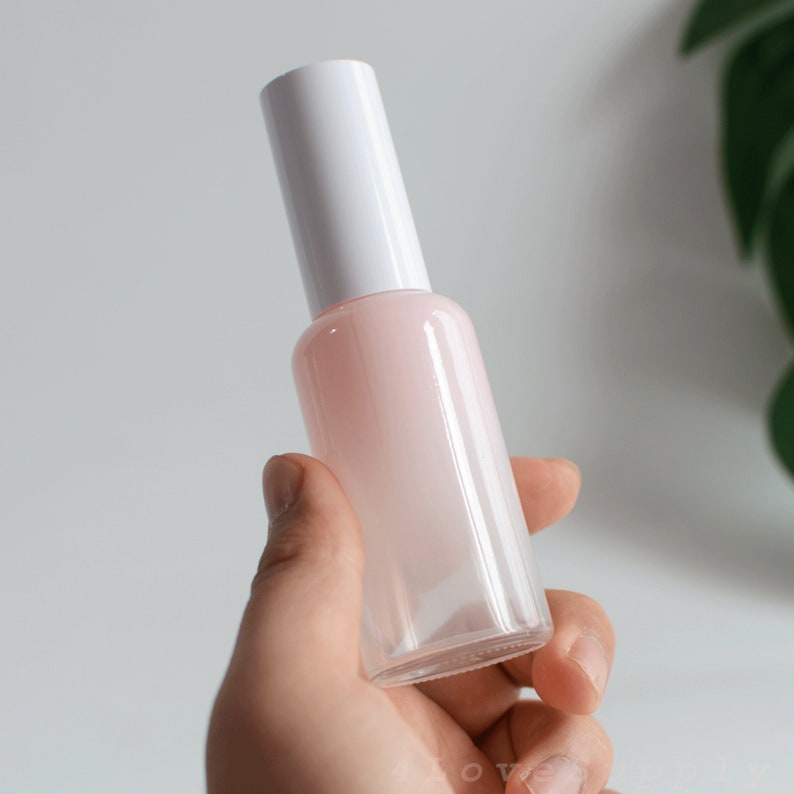 May include: A clear glass bottle with a white cap and a light pink liquid inside. The bottle is being held in a hand.
