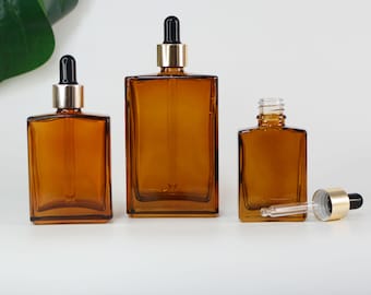 May include: Three amber glass dropper bottles of varying sizes. Each bottle has a gold-colored collar and a black dropper top. One bottle has the dropper removed. The bottles are rectangular with rounded corners.