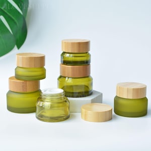 May include: A collection of frosted olive green glass cosmetic jars with light-colored bamboo lids. The set includes several stacked and individual jars, one of which is open to reveal a white inner seal, arranged on a white surface.