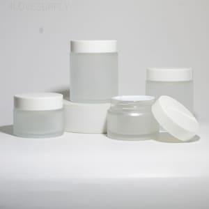 Op de afbeelding: Verschillende matglazen potten met witte deksels staan op een wit oppervlak. Eén pot heeft het deksel eraf, waardoor de inhoud zichtbaar is. De potten variëren in grootte, wat wijst op een set voor cosmetica- of huidverzorgingsproducten. De tekst "4LOVESUPPLY" is zichtbaar.