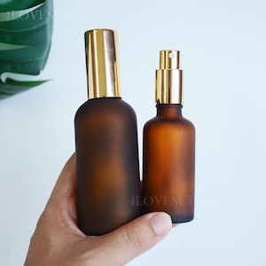 May include: Two amber-coloured frosted glass bottles with gold-toned spray pump caps. The taller bottle features a smooth cylindrical body, while the shorter bottle has a slightly more compact design. Both containers are opaque and matte in texture.
