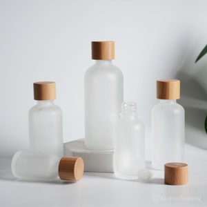 May include: Five frosted glass bottles with bamboo lids. The bottles are various sizes and are arranged on a white surface.