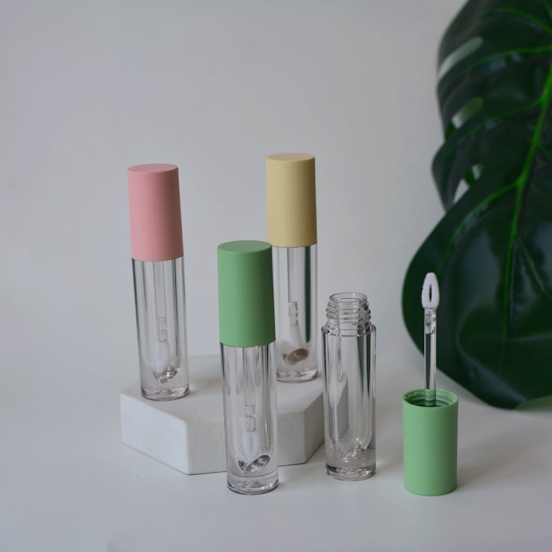 May include: Four clear lip gloss tubes with different colored caps. The caps are pink, yellow, green, and green. The tubes are on a white hexagonal surface.