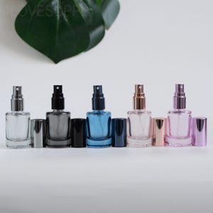 May include: A row of five glass spray bottles in various colors: clear, gray, blue, rose gold, and pink. Each bottle has a matching cap and a silver or rose gold spray nozzle. The bottles are rectangular and sit on a white surface.