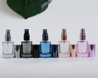 May include: A row of five glass spray bottles in various colors: clear, gray, blue, rose gold, and pink. Each bottle has a matching cap and a silver or rose gold spray nozzle. The bottles are rectangular and sit on a white surface.