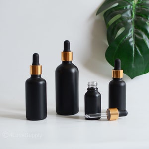 Könnte beinhalten: Vier schwarze Glas-Tropfflaschen mit goldfarbenen Kragen und schwarzen Tropfern. Eine Flasche ist geöffnet und zeigt die Glastropfpipette. Die Flaschen sind auf einer weißen Oberfläche angeordnet, mit einem großen grünen Blatt im Hintergrund.