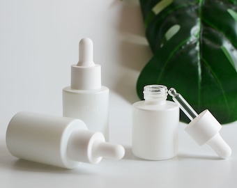 May include: Three white frosted glass bottles with dropper tops. The bottles are on a white surface with a green leaf in the background.