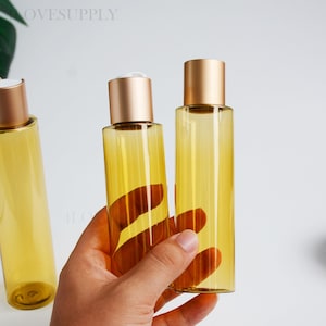 Puede incluir: Tres botellas cilíndricas de plástico de color ámbar con tapas de rosca de oro metálico. Las botellas son translúcidas, permitiendo que la luz pase a través del material teñido de amarillo. Cada botella tiene un diseño liso.