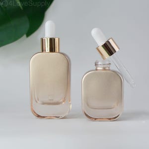 Könnte beinhalten: Zwei Kosmetikflaschen aus Glas mit goldfarbenen Deckeln und weißen Pipetten. Eine Flasche ist geöffnet, mit der Pipette teilweise eingesetzt. Die Flaschen haben eine abgerundete rechteckige Form und ein mattiertes, hellbraunes Aussehen.