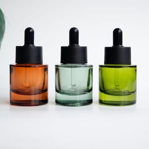 Könnte beinhalten: Drei Glas-Tropfflaschen in Orange, Grün und Olivgrün. Jede Flasche hat einen schwarzen Deckel und eine Glastropfpipette. Die Flaschen sind zylindrisch und stehen auf einer weißen Oberfläche. Wahrscheinlich für ätherische Öle oder Seren.