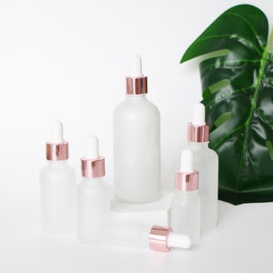 May include: A collection of frosted glass dropper bottles with rose gold collars and white droppers. The bottles vary in size and are arranged on a white surface. A large green leaf is in the background.