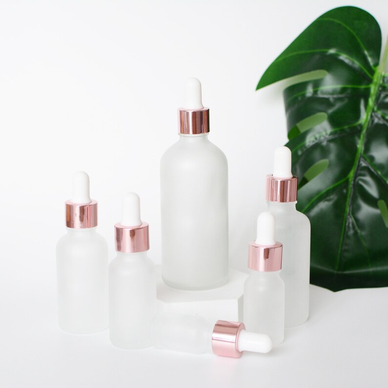 May include: A collection of frosted glass dropper bottles with rose gold collars and white droppers. The bottles vary in size and are arranged on a white surface. A large green leaf is in the background.