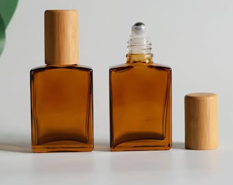 Könnte beinhalten: Zwei rechteckige bernsteinfarbene Glasflaschen mit Bambusdeckeln. Eine Flasche ist verschlossen, während die andere offen ist und einen Roll-on-Einsatz aus klarem Kunststoff zeigt. Die Flaschen haben einen flachen Boden und eine glatte, braune Oberfläche.