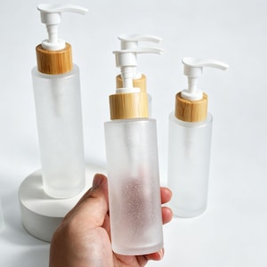 Puede incluir: Tres botellas cilíndricas de vidrio esmerilado con dispensadores de bomba de plástico blanco y collares de bambú claro. Las botellas tienen un acabado translúcido y texturizado. Una botella se sostiene en primer plano, dos detrás.