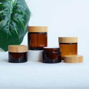 以下が含まれることがあります： 竹製の蓋が付いた琥珀色のガラス瓶がいくつか表示されています。瓶はサイズと形状が異なり、中央に小さな開いた瓶があります。蓋は薄茶色で、瓶は白い表面に配置されています。